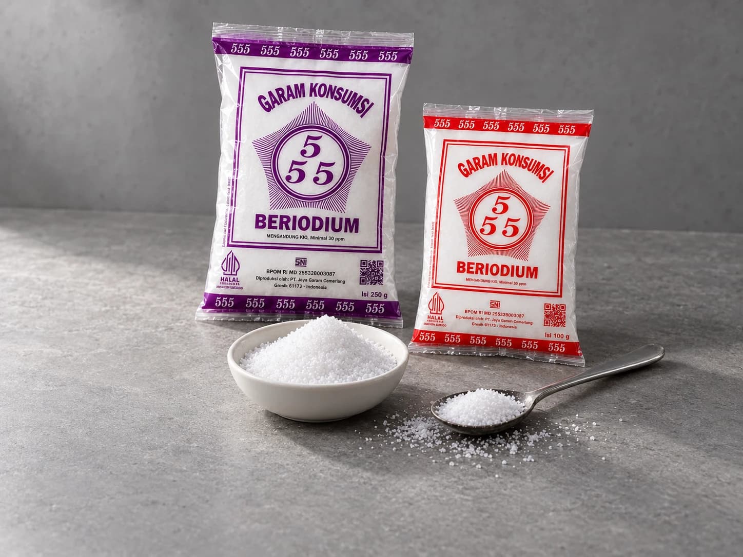 Kemasan Cap 555 ungu 250 gram dan merah 100 gram bersama sampel garam dalam mangkuk putih dan sendok logam di meja abu-abu.